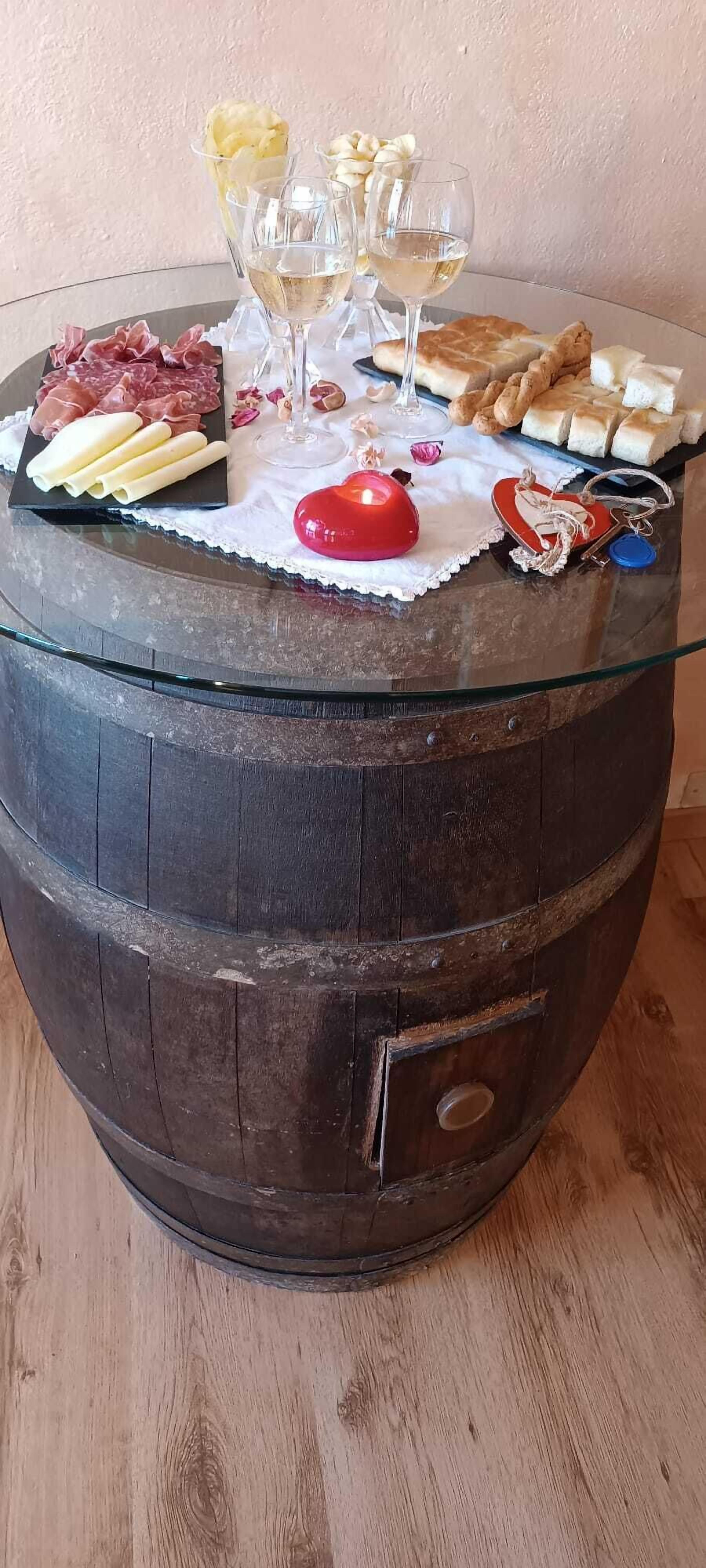 Una mesa rústica de madera con cubierta de vidrio, decorada con una variedad de quesos, embutidos y pequeños pasteles.