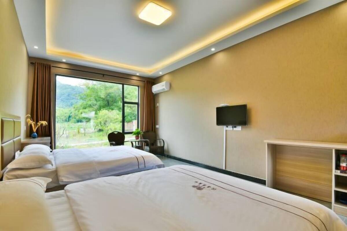 A hotel room with two beds, a TV, a desk, and a window with a view of greenery.
