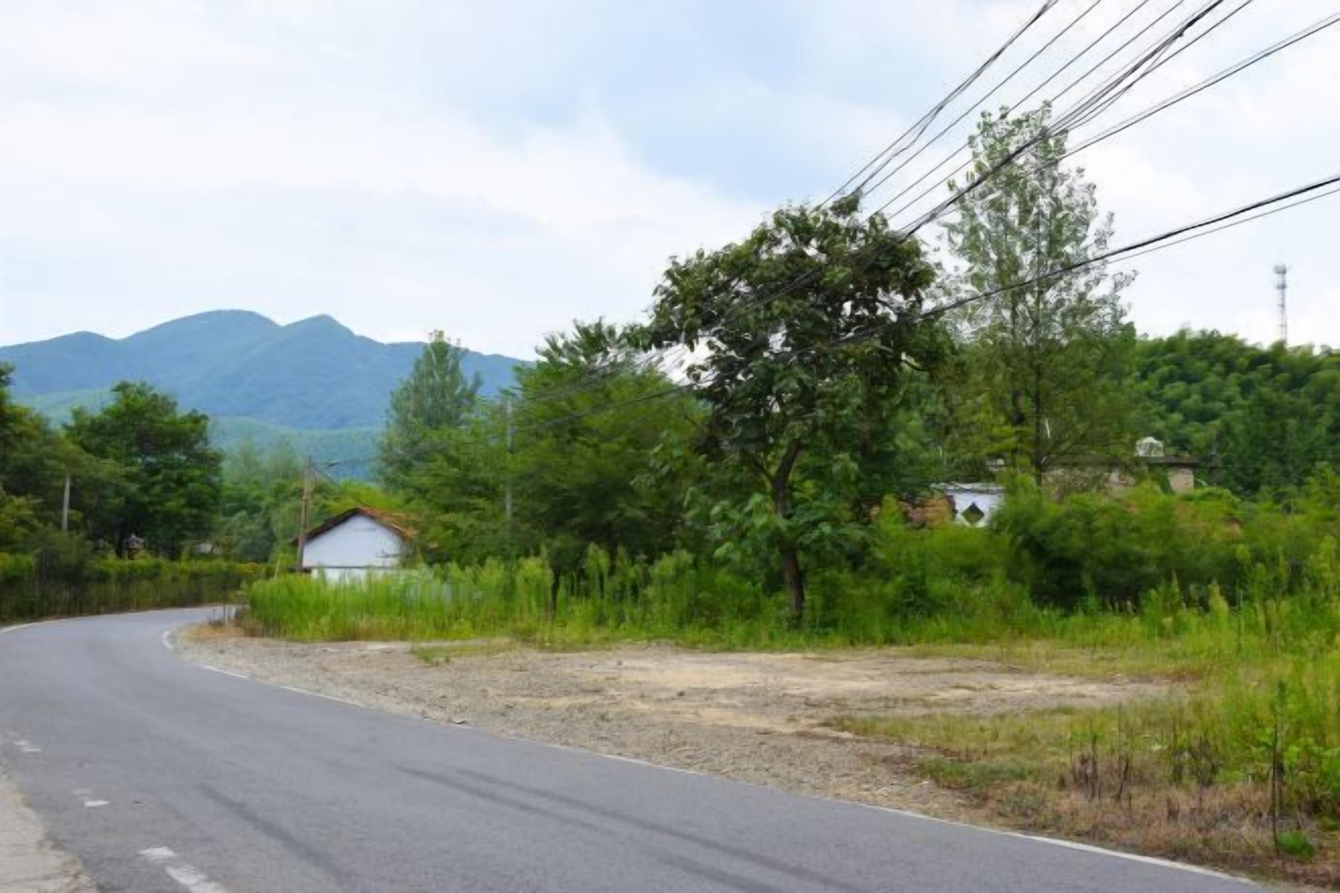 Eine ländliche Straße mit einem Haus links, umgeben von Grünflächen und Hügeln im Hintergrund.