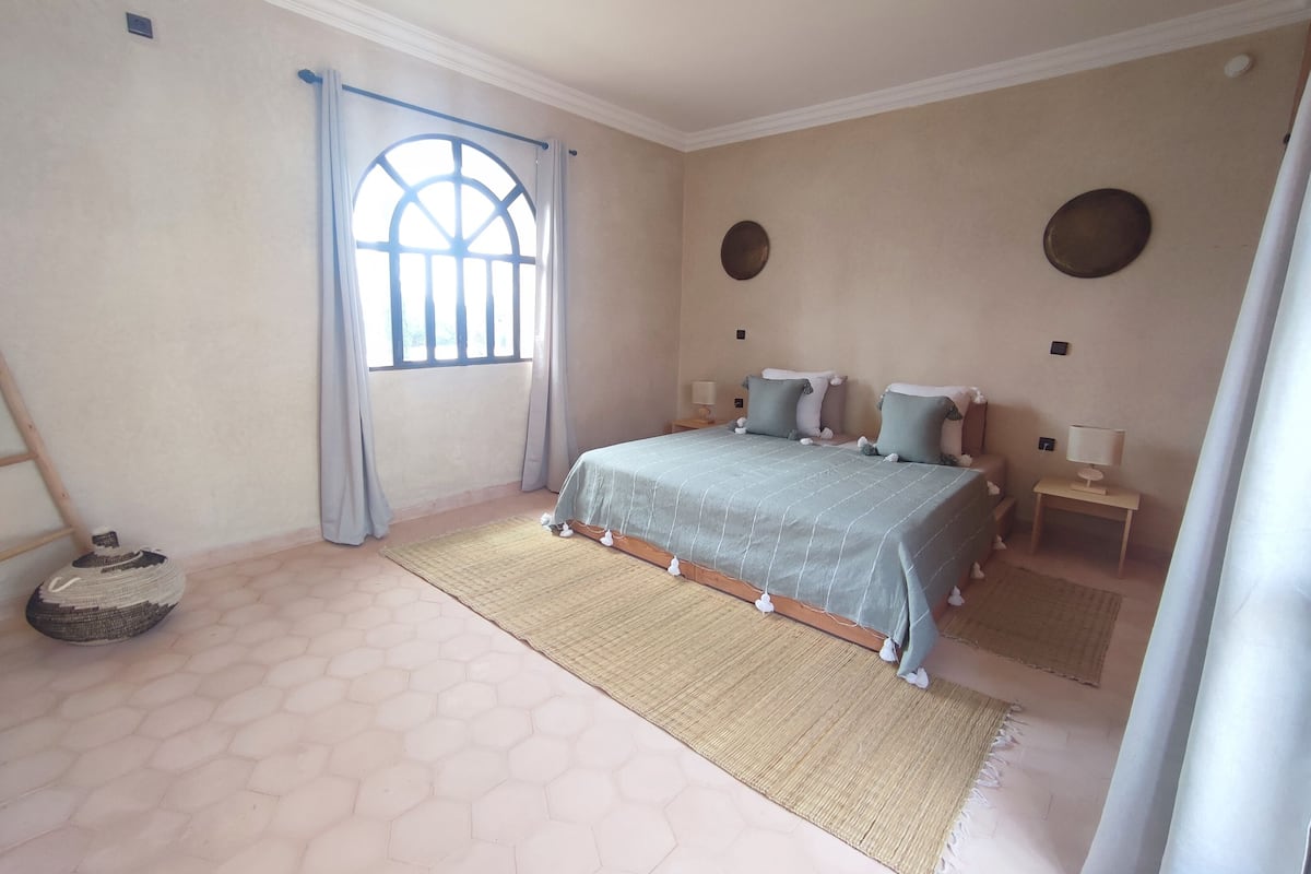 A bedroom with a large bed, two bedside tables with lamps, a window with curtains, and a hexagonal floor pattern.
