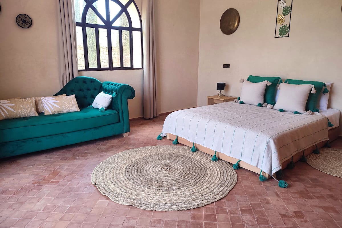 A room with a green sofa, a bed with a white and green bedspread, a woven rug, and a stone wall.