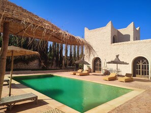 A pool area with lounge chairs, a thatched umbrella, and a stone building with arched windows. - Riad Bini- Ddik (Aguerd)