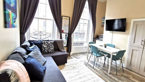 A modern living room with a sofa, dining table, and chairs. There is a TV mounted on the wall and a window with curtains. - Great Central Location 1BED APT Off OConnell ST (Dublin 1)