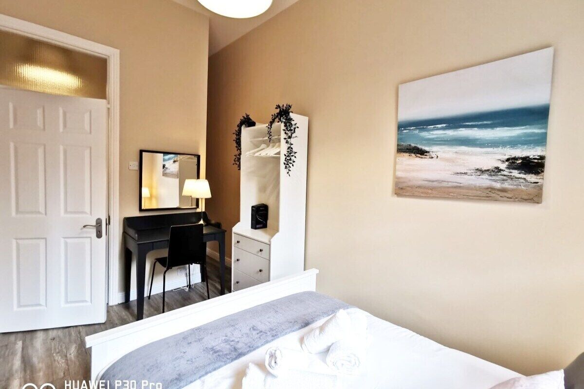 Quarto de hotel com uma cama, uma escrivaninha com cadeira, cómoda, espelho e um quadro de uma praia.