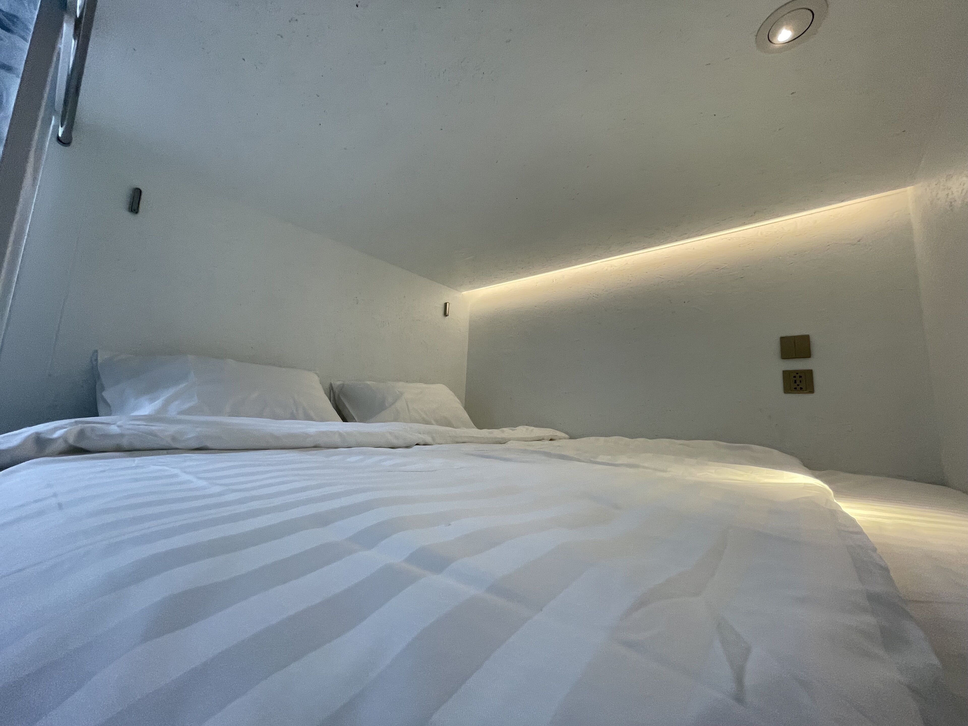A small, dimly lit room with a single bed, a ceiling light, and a wall socket.