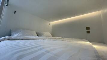 A small, dimly lit room with a single bed, a ceiling light, and a wall socket.