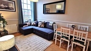 Ein modernes Wohnzimmer mit einem dunklen Sofa, einem Esstisch mit weißen Stühlen, einem großen Spiegel und einem Fenster mit Vorhängen.