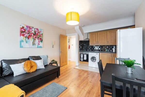 Um apartamento moderno com sala de estar, um sofá preto, mesa de jantar e cozinha com armários de madeira e máquina de lavar.