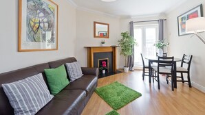 Een woonkamer met een bruine leren bank, groene en gestreepte kussens, een groen vloerkleed, een open haard, een eettafel met stoelen en een groot abstract schilderij aan de muur.