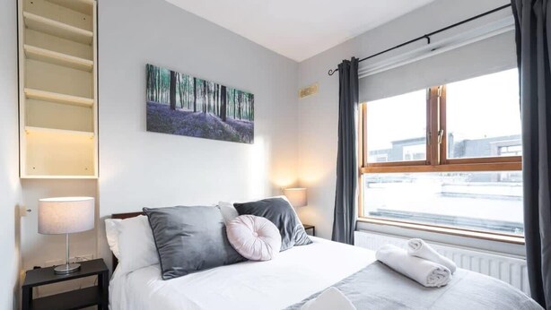 A hotel room with a bed, a painting on the wall, a window with curtains, and a nightstand with a lamp. - GREAT NEIGHBORHOOD SUPER 2 bed COTTAGE SUIT FAMILY (Dublin 8)