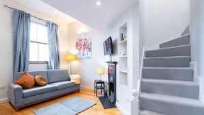 A living room with a grey sofa, a small table, a fireplace, and a staircase. - GREAT NEIGHBORHOOD SUPER 2 bed COTTAGE SUIT FAMILY (Dublin 8)