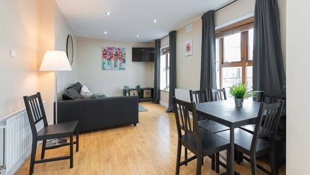 A modern living room with a sofa, dining table, and chairs. There is a TV mounted on the wall, a window with curtains, and a radiator on the floor. - Great City CENTRE LOCATION SPACIOUS 2 BED APT (Dublin 1)