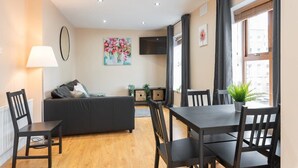 A modern living room with a sofa, dining table, and chairs, a TV, and a large window with curtains. - Great City CENTRE LOCATION SPACIOUS 2 BED APT (Dublin 1)