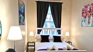 Ein Hotelzimmer mit Bett, zwei Nachttischlampen, einem Fenster mit Vorhängen und Bildern an den Wänden.