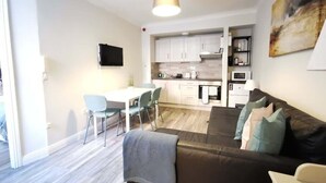 Een moderne woonkamer met een bank, een eethoek met stoelen, een keuken met kasten, en een aan de muur bevestigde televisie.