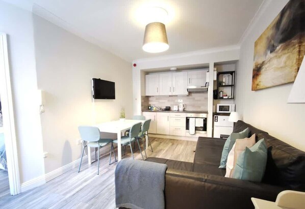 A modern living room with a sofa, a dining area with chairs, a kitchen with cabinets, and a TV mounted on the wall.