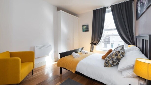Moderni hotellihuone, jossa on sänky, keltainen nojatuoli, valkoinen vaatekaappi ja ikkuna, jossa on verhot.