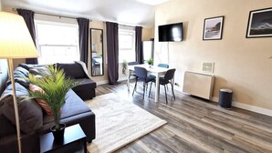 En moderne stue med sofa, sofabord, lampe, spiseområde med stole, et fjernsyn ophængt på væggen og et vindue med gardiner.