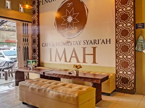 Un espace d’accueil avec un bureau en bois, un banc rembourré et un mur sur lequel on peut lire : « Café & hébergement Syari’ah IMAH ».
