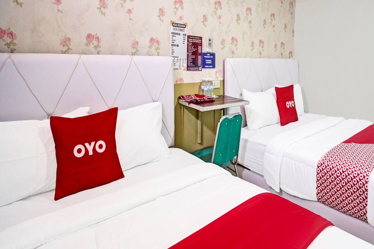 Une chambre d’hôtel avec deux lits, chacun étant équipé de draps blancs et d’oreillers rouges. On y trouve une petite table avec une chaise, une bouteille d’eau et un menu affiché au mur.