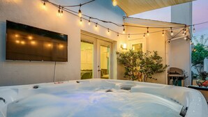 A hot tub with a flat-screen TV mounted on the wall, string lights, and a potted plant.
