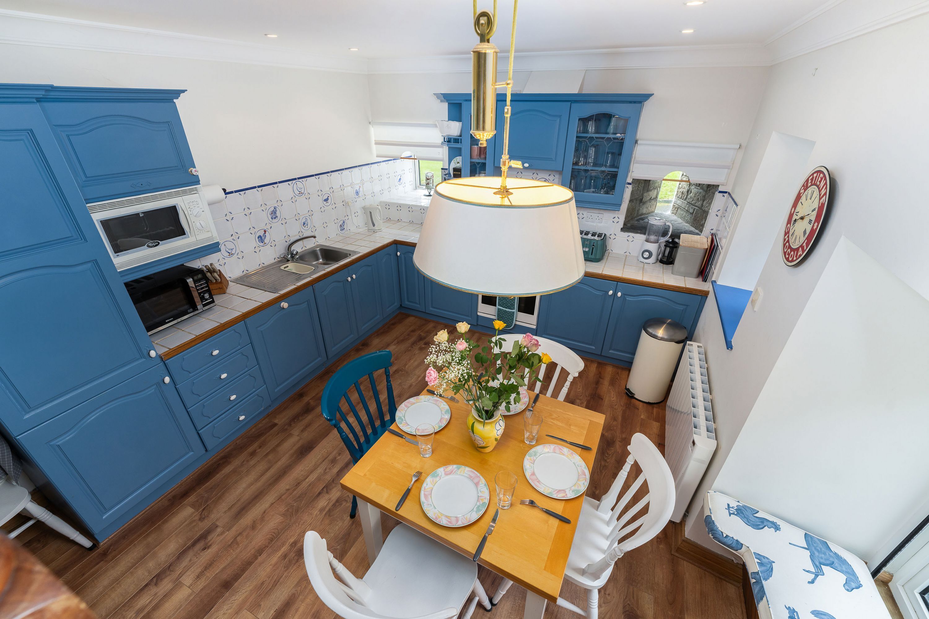 Une cuisine moderne avec des meubles bleus, une table à manger en bois et des chaises blanches, ainsi qu’un luminaire suspendu.
