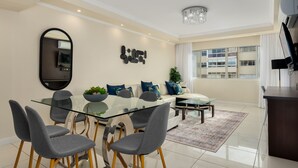 A modern living room with a glass dining table, grey chairs, a sofa with blue cushions, a chandelier, and a large window with a view of a building. - New Cumberland 405 (Cape Town)