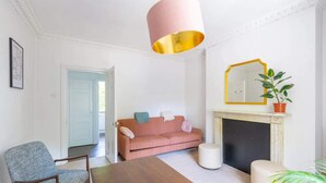 Ett vardagsrum med ett träbord, en rosa soffa, en blå fåtölj, en vit pall, en öppen spis med en guldfärgad spegelram och en krukväxt.