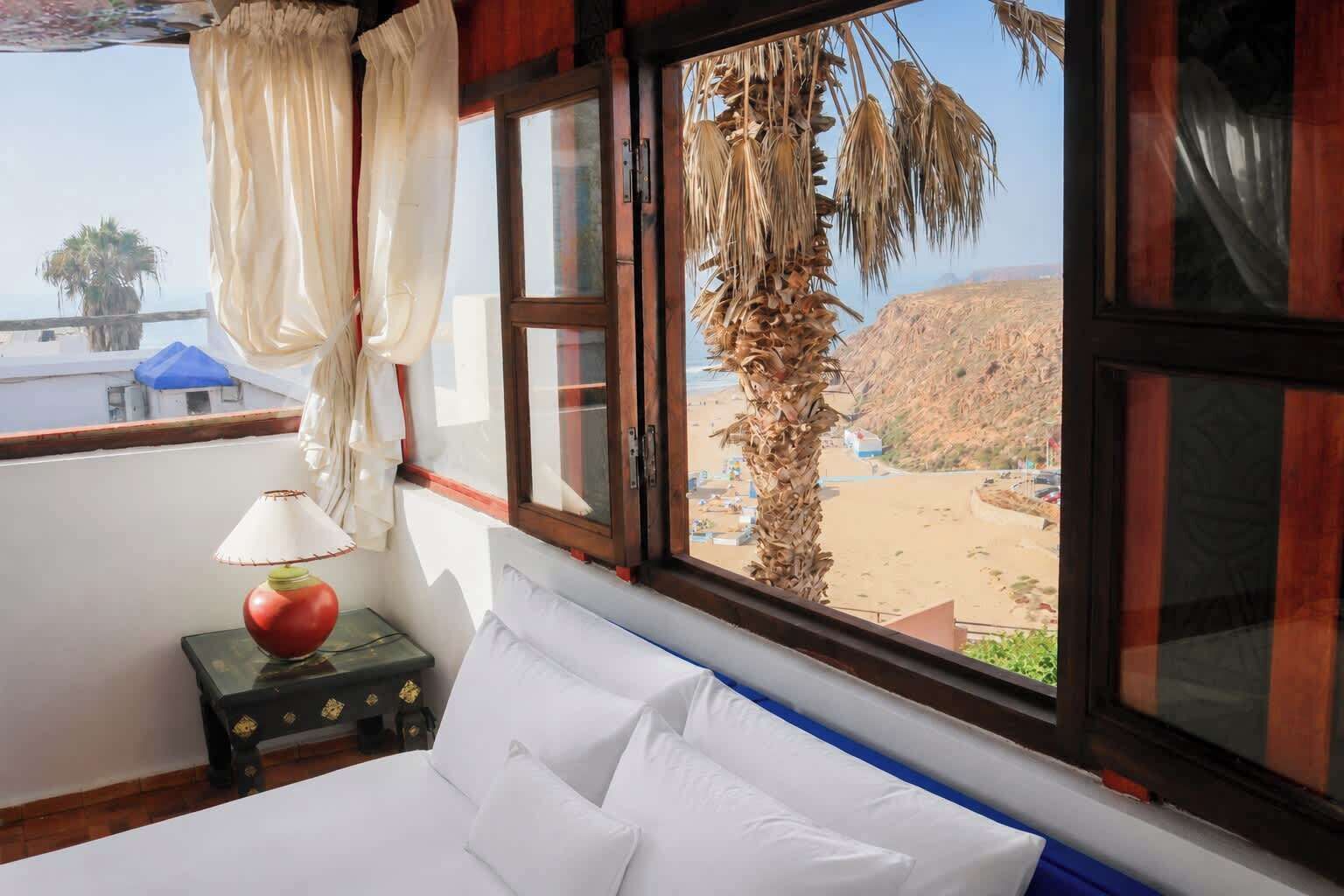 Un dormitorio con cama, una mesita de noche con lámpara y vistas a una playa y una palmera a través de amplios ventanales.