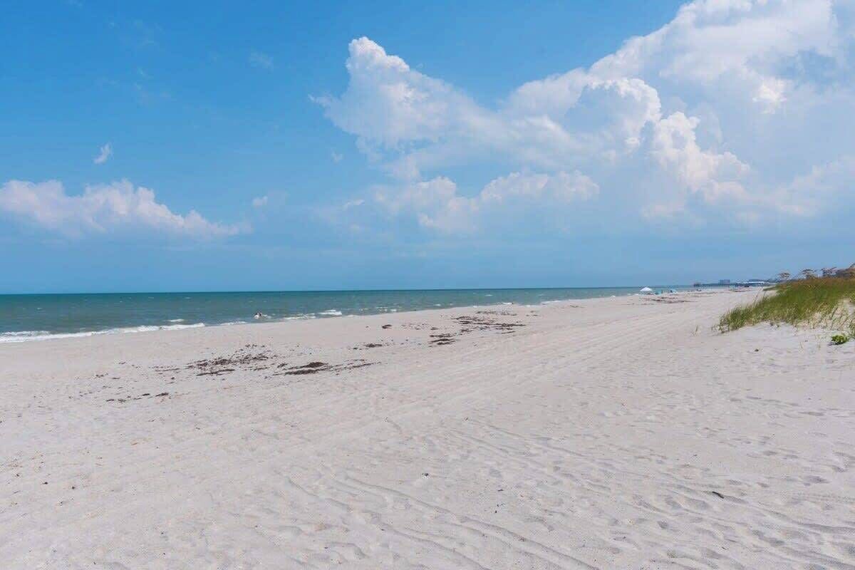 A wide sandy beach with gentle waves, a clear sky, and distant dunes.