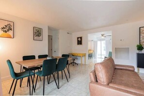 A modern living room with a brown leather sofa, a dining area with a wooden table and teal chairs, and a kitchen area with a yellow countertop. - Fillmore Unit 4 | 4BR/2BA (Cape Canaveral)