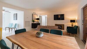 A modern living room with a wooden dining table, green chairs, a fireplace, and a flat-screen TV mounted on the wall.