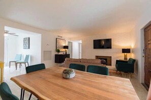 Een moderne woonkamer met een houten eettafel, groene stoelen, een open haard en een flatscreen televisie aan de muur.