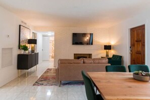 A modern living room with a fireplace, a television mounted on the wall, and a dining area with a wooden table and chairs. - Pool condo w/ patio | steps to the beach (Cape Canaveral)