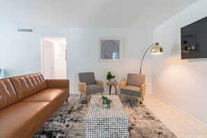 A living room with a brown leather sofa, two armchairs, a coffee table with a patterned rug, a floor lamp, and a framed artwork on the wall. - Fillmore Unit #5 | 2BR/1BA (Cape Canaveral)