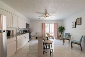 A kitchen with white cabinets, a central island with stools, a ceiling fan, and a dining area with chairs and a table. - Fillmore Unit #5 | 2BR/1BA (Cape Canaveral)
