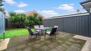 A patio with a table and chairs, a grassy lawn, a brick building, and a metal fence. - Mellow&Modernized Townhouse-3Beds-Quiet-Anzac Hwy (Plympton)