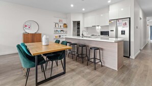 A modern kitchen with white cabinetry, a central island with stools, and a dining area with a wooden table and green chairs. - Mellow&Modernized Townhouse-3Beds-Quiet-Anzac Hwy (Plympton)