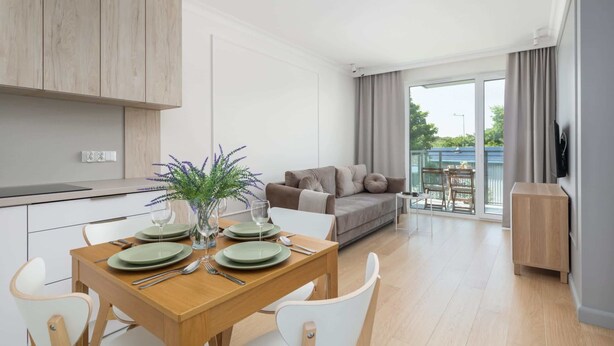 Una cocina moderna con armarios blancos, una mesa de comedor con platos y cubiertos, y vistas al área de estar con un sofá y un balcón.