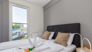 Una cama bien hecha con sábanas blancas, un cabecero gris y almohadas color beige. Se ve una mesita de noche con una lámpara blanca.