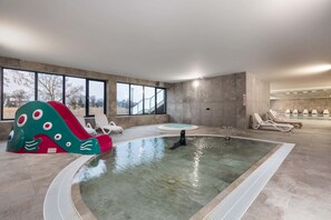 Une piscine intérieure avec un toboggan, des chaises longues et une vue sur l’extérieur grâce à de grandes fenêtres.