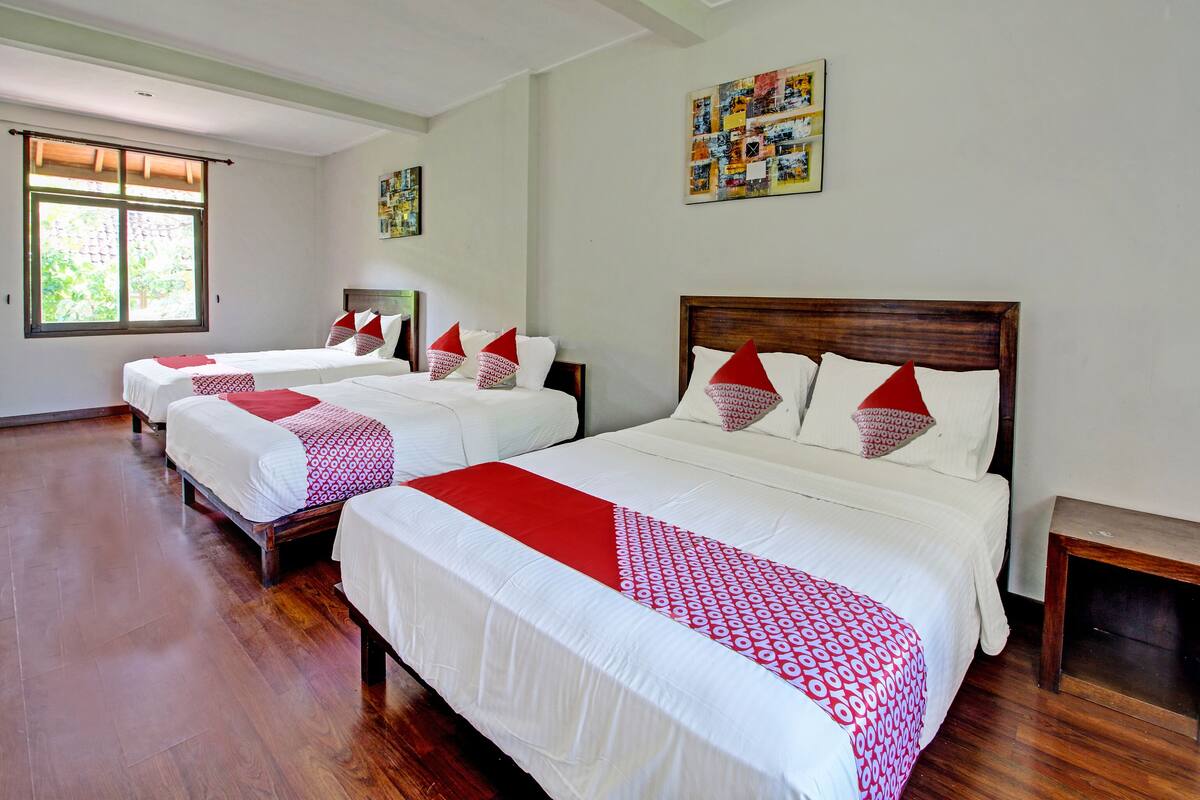 A hotel room with three beds, each with white bedding and red accents, wooden headboards, and a large window providing natural light.
