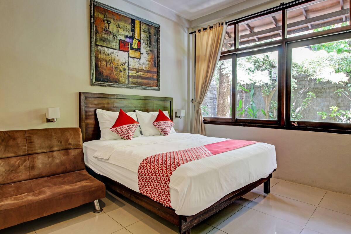 A hotel room with a bed, a brown sofa, a painting on the wall, and a window with curtains.