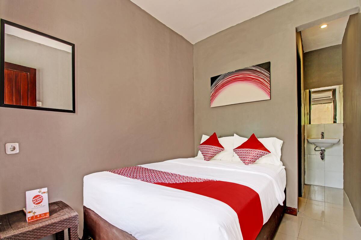 A hotel room with a bed, a nightstand, a mirror, a painting, and a bathroom visible through an open door.