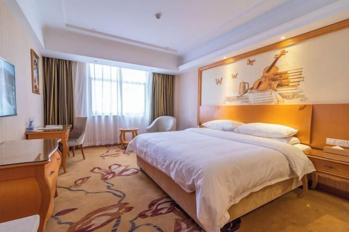 A hotel room with a bed, a desk, a chair, a window with curtains, and a wall with a guitar and books mural.