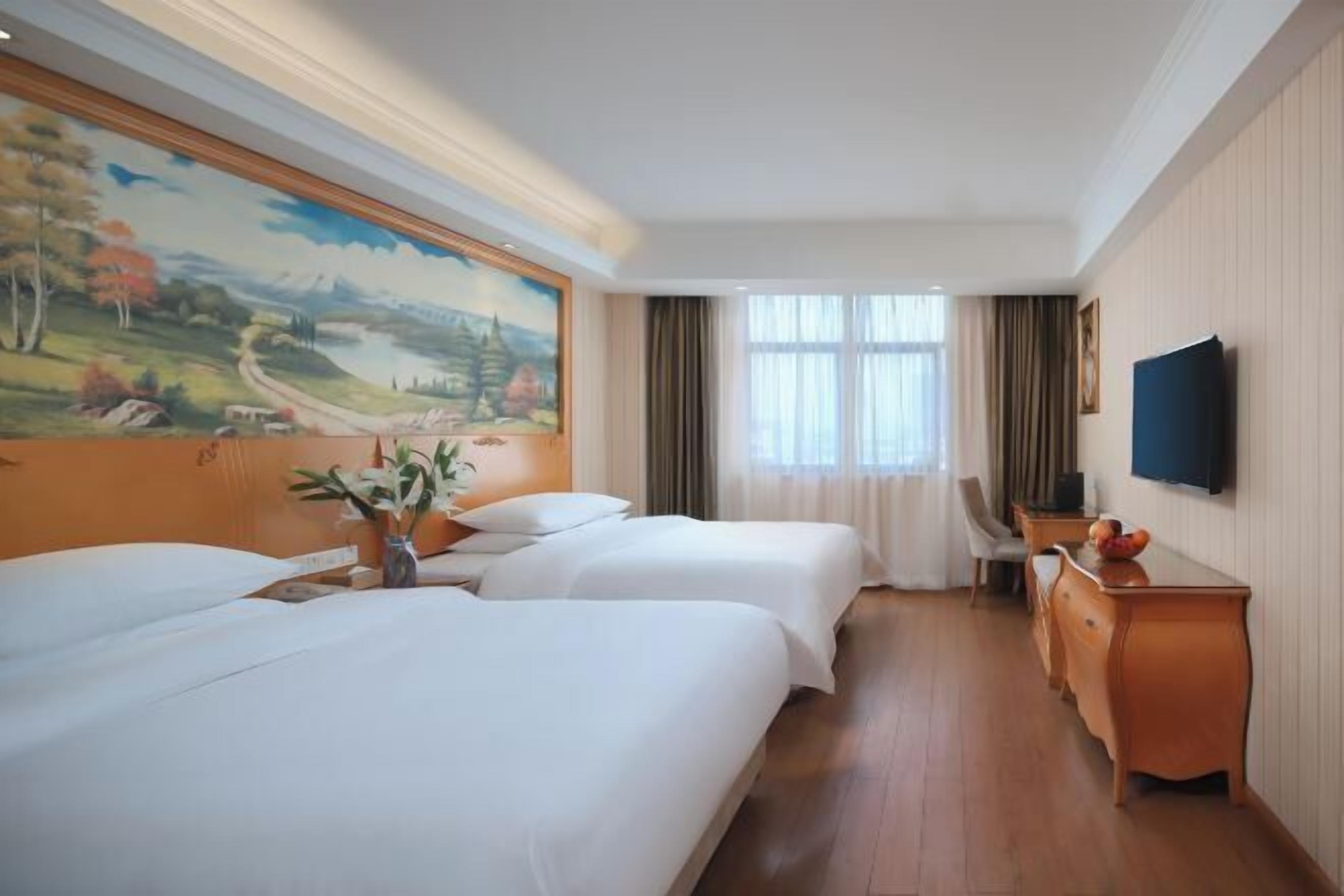 Habitación de hotel con dos camas, un aparador de madera, un escritorio, un televisor y un cuadro grande de paisaje en la pared.