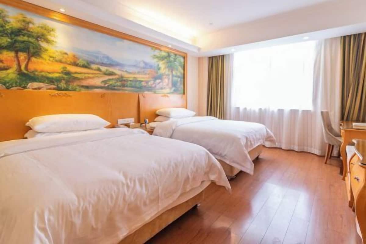 A hotel room with two beds, a painting on the wall, a window with curtains, and a wooden floor.