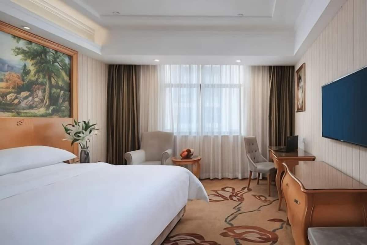 A hotel room with a large bed, a desk, a chair, a TV, and a painting on the wall.