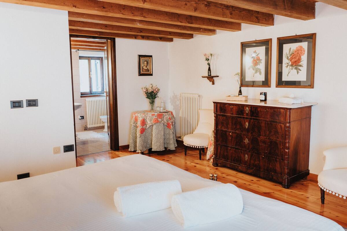 Un dormitorio con piso de madera, una cama con ropa de cama blanca, un aparador de madera y una silla.