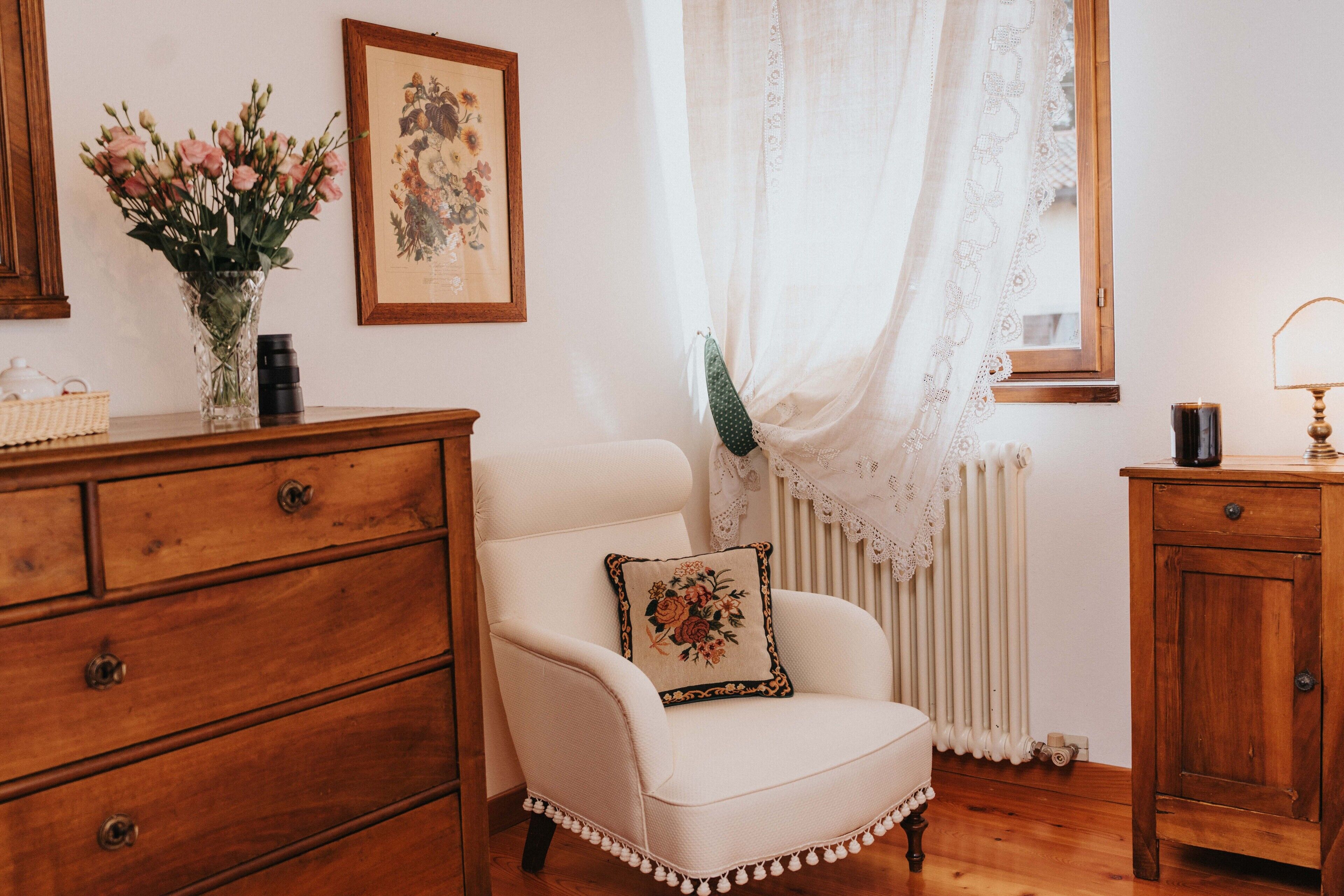 Una habitación con un aparador de madera, un sillón blanco con un cojín floral, un jarrón con flores, un cuadro en la pared y un radiador.